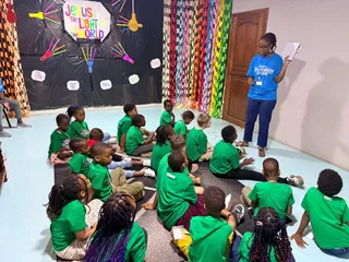 Children in green shirts sitting on the floor as a teacher reads to them