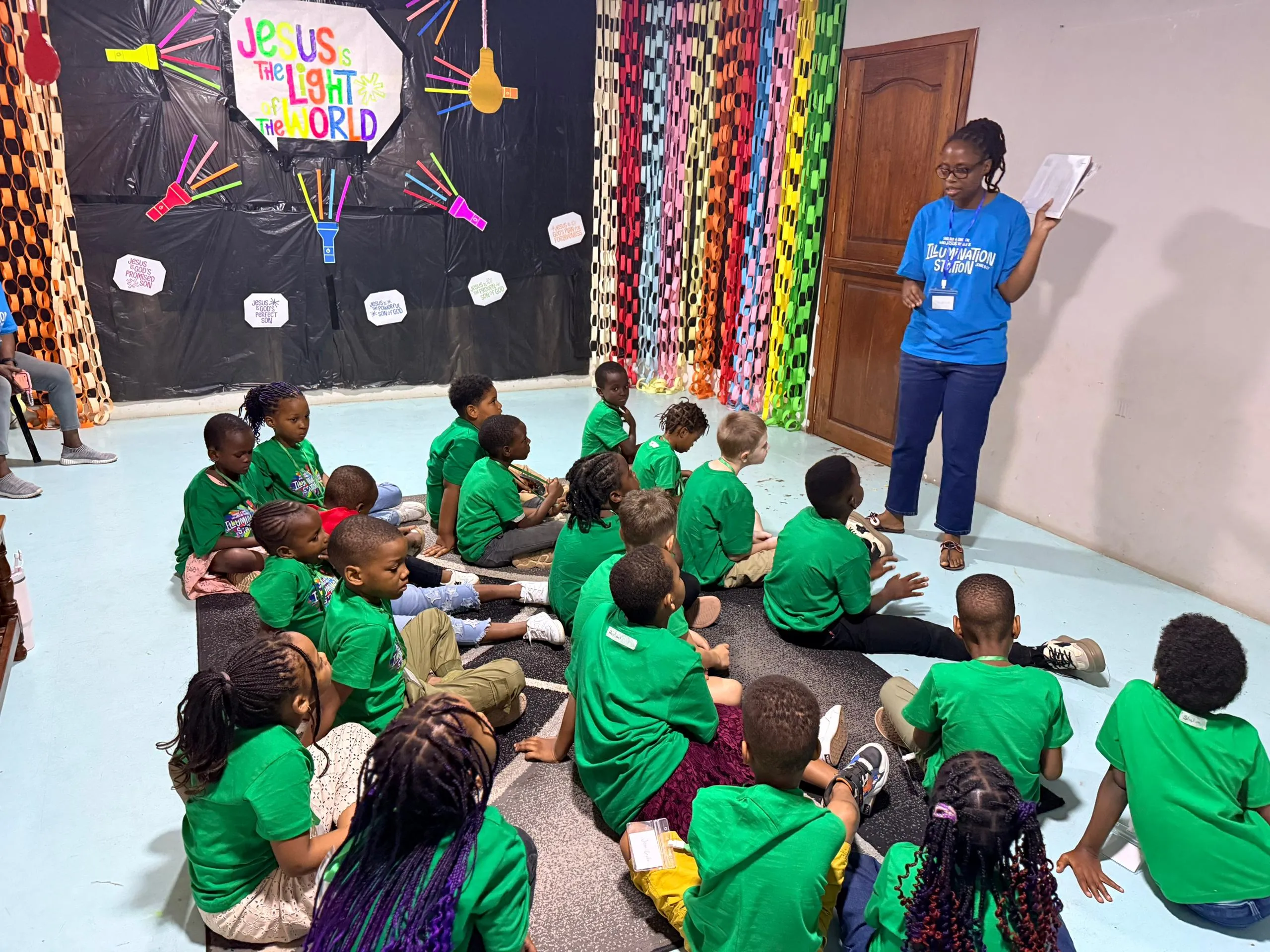 Children in green shirts sitting on the floor as a teacher reads to them